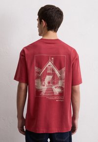 Rotes Baumwoll-T-Shirt mit einem weißen Skizze eines Hauses auf dem Rücken, mit Text darunter. Bequeme Passform und kurze Ärmel.