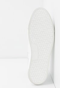 Baskets blanches avec une semelle en caoutchouc texturée à motif circulaire, un dessus lisse et un design minimaliste sur fond blanc.