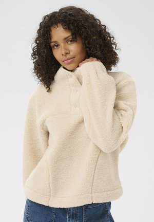 Mlajša ženska s kodrastimi lasmi, oblečena v kremasto sherpa pulover in modre kavbojke, drži ovratnik z eno roko.