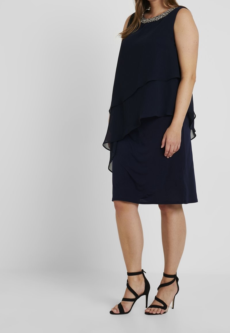 Marinblå lagerklänning med rund halsringning och pärlaccenter. I kombination med svarta, remmar med hög klack. Slätt, flödande tyg.