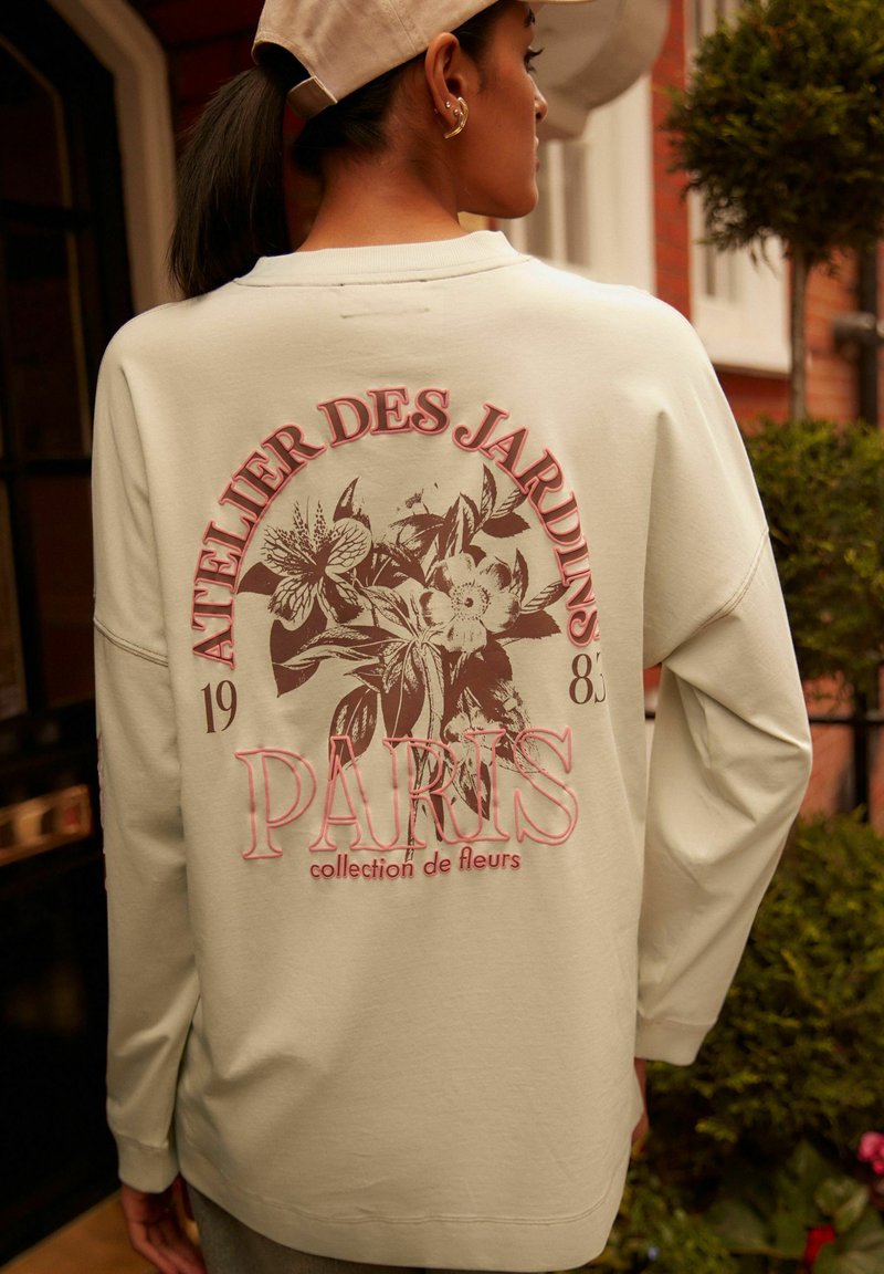 Világos bézs pulóver, rózsaszín és fekete virágos grafikai nyomattal, az "ATELIER DES JARDINS 1983 PARIS collection de fleurs" felirattal.