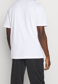 Camiseta blanca de algodón con corte relajado, mangas cortas, diseño liso y cuello redondo; vista trasera con pantalones oscuros.