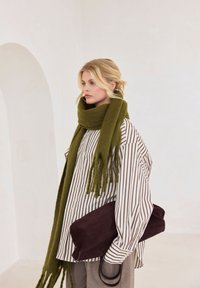 Écharpe vert olive à franges, chemise rayée blanche et marron, sac en daim marron foncé, et pantalon gris. Design simple et moderne.