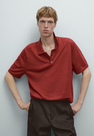 Rød kortærmet polo-shirt med knapkrave, lavet af blødt, tekstureret stof, parret med brune bukser, stående mod en lys væg.