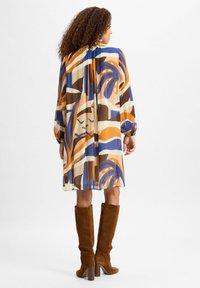 Fließendes Kleid in Creme, Braun, Blau und orangefarbenem abstraktem Muster; lange Ärmel; geraffter Ausschnitt; kombiniert mit kniehohen, braunen Wildlederstiefeln.