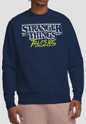Mann trägt marineblaue Sweatshirt mit weißem und grünem Schriftzug "Stranger Things Tales from '85" auf der Vorderseite.