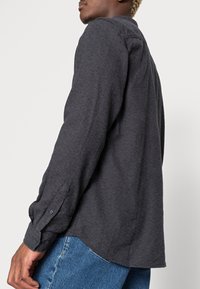 Długa rękaw koszula w kolorze ciemnoszarym, wykonana z teksturowanej tkaniny, z mankietami zapinanymi na guziki i zaokrąglonym dołem, noszona z niebieskimi dżinsami.