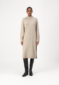 Beige neulemekko, jossa on korkea kaulus, ribbireunat ja sivuaukot, joita täydentävät tyylikkäät mustat polvipituiset saappaat. Sujuva, pehmeä rakenne.