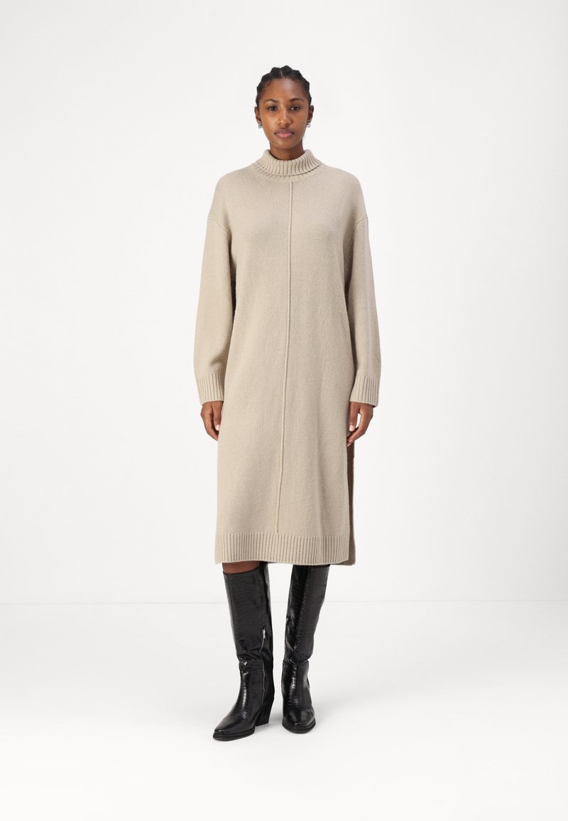 Beige neulemekko, jossa on korkea kaulus, ribbireunat ja sivuaukot, joita täydentävät tyylikkäät mustat polvipituiset saappaat. Sujuva, pehmeä rakenne.