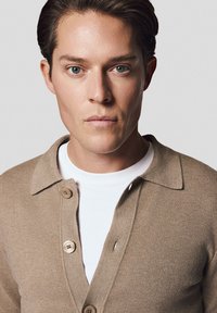 Beżowy sweter z kołnierzykiem, wykonany z teksturowanej tkaniny i zapinany na drewniane guziki, noszony na białej koszulce. Dopasowany krój.