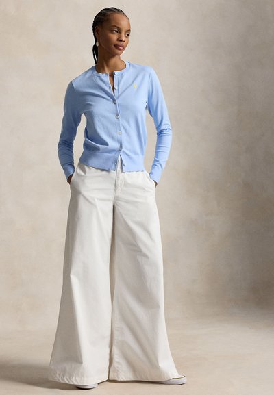 Frau trägt eine hellblaue, geknöpfte Strickjacke und weiße, weit geschnittene Hosen, steht mit den Händen in den Taschen vor einem neutralen Hintergrund.