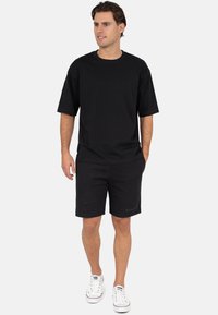 T-shirt en coton noir à manches courtes, associé à un short noir avec un petit logo. Porté avec des baskets blanches, debout sur un fond uni.