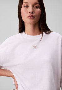 Joven mujer con el pelo largo y oscuro que lleva una camiseta blanca lisa y grande con un pequeño logo de Calvin Klein en el pecho, mano en la cadera.