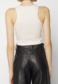 Kvinna klädd i en vit ärmlös ribbad crop top och högmidjade svarta läderbyxor, sedd bakifrån mot en neutral bakgrund.