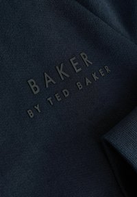 Jūras spēku audums ar gludu tekstūru, kurā ir iespiests zīmols: "BAKER" un "BY TED BAKER" kontrastējošā melnā krāsā.