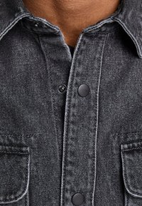 Nahaufnahme einer dunkelgrauen Jeansjacke mit schwarzen Druckknöpfen und zwei vorderen Brusttaschen, die am Kragen teilweise offen ist.