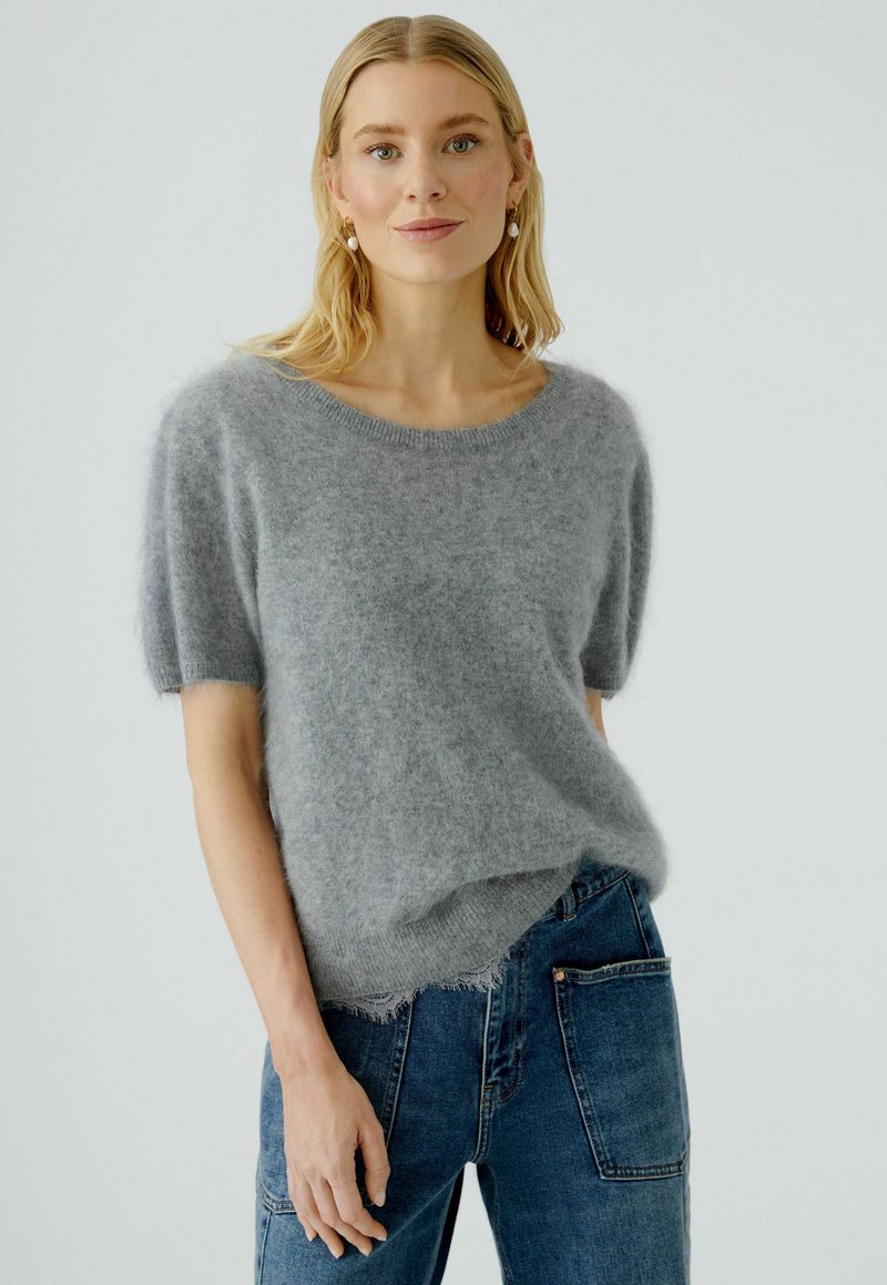 Hellgrauer, kurzärmeliger Pullover aus flauschigem Material, mit rundem Hals und leicht ausgefranstem Saum, kombiniert mit blauen Jeans.