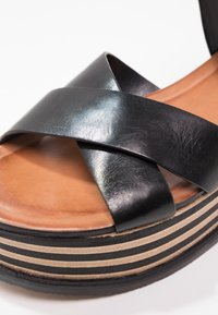 Black leather platform sandal with crossed straps, tan insole, and a striped cork and black rubber sole. Smooth texture, modern design.
