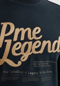 Mornarica dukserica s istaknutim zlatnim vezom "Pme Legend" i tekstom koji opisuje povijest zrakoplovstva. Mekani materijal s okruglim izrezom.