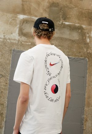 Person, die ein weißes T-Shirt mit kreisförmigem schwarzem Text, rotem und schwarzem Yin-Yang-Symbol sowie einem roten Schwung auf dem Rücken trägt, steht vor einer strukturierten Wand.