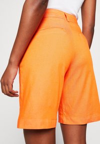 Žena v jasně oranžových šortkách s vysokým pasem a bílém topu bez rukávů, zobrazena zezadu ve výšce pasu a stehen.