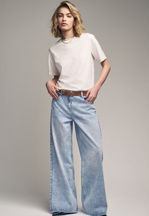 T-shirt en coton beige clair associé à un jean en denim clair à jambes larges. Une ceinture en cuir marron est visible, créant une silhouette décontractée et relaxante.