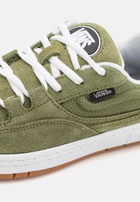 Baskets en daim vert avec lacets blancs et logo Vans noir. Elles présentent une semelle intermédiaire blanche et une semelle extérieure en caoutchouc naturel. Détails texturés sur la tige.