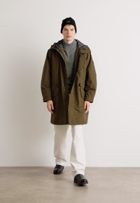 Casaco parka verde-oliva com capuz, dois bolsos frontais e cordão. Combinado com um suéter cinza de gola alta e calças brancas, apresentando sapatos pretos.