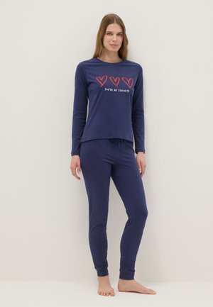 Frau steht barfuß und trägt dunkelblaue Pyjamas mit langärmligem Oberteil und Hose, auf dem Oberteil drei rote Herzen und der Schriftzug "Du bist mein Lieblingsmensch".