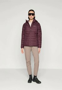 Giacca a piumino bordeaux con chiusura a zip e cappuccio, caratterizzata da cuciture orizzontali. Indossata con pantaloni beige e scarpe nere. Occhiali da sole scuri.