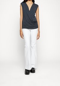 Dunkelgraues, drapiertes V-Ausschnitt-Top mit kurzen Ärmeln, kombiniert mit weißen, ausgestellten Hosen. Das Model trägt schwarze Schuhe. Einfaches, klares Design.