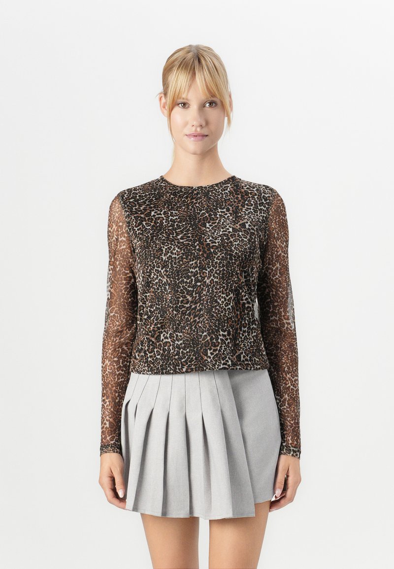 Woman wearing a sheer long-sleeve leopard print top and a light gray pleated mini skirt, standing against a plain background.