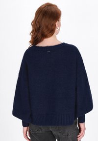 Granatowy, oversize sweter o miękkiej, puszystej fakturze. Posiada opadające ramiona, ribbrowany dół oraz subtelna metkę z logo na plecach.