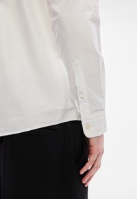 Λευκό πουκάμισο με μακριά μανίκια και κουμπιά στα μανικέτια, με λεία υφή και εφαρμοστή γραμμή, μερικώς ε�μφανιζόμενο πάνω σε μαύρο παντελόνι.