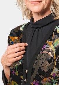 Schwarze Strickjacke mit Blumenmuster, farbigen Akzenten, samtiger Textur und goldenen Knöpfen. Schwarzes Oberteil mit hohem Kragen darunter. Nahaufnahme des Ausschnitts.