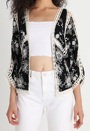 Mujer con un kimono negro abierto al frente con bordado floral blanco, sobre una camiseta corta blanca y pantalones blancos de talle alto.