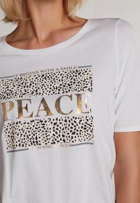 Witte T-shirt met korte mouwen met gouden folieletters. Het ontwerp bevat de woorden "PEACE" en een gespikkeld patroon in zwart op een beige achtergrond.