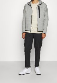 Šedá mikina na zip s černými doplňky, doplněná černými cargo kalhotami. Outfit je dokončen bílými teniskami s černým lemováním.