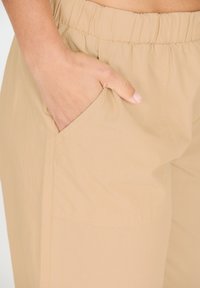 Beige housut elastisella vyötäröllä, jossa on sileä pinta ja sivutaskut, yksinkertainen, minimalistinen muotoilu ilman kuvioita tai koristeita.