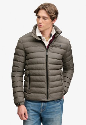 Young man wearing a zipped brown checkered puffer jacket with hands in pockets, looking slightly downward to his left.