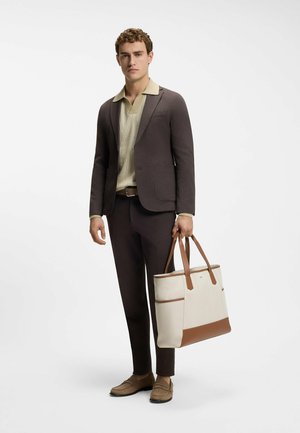 Homme portant un costume marron foncé et une chemise beige, tenant un grand sac fourre-tout beige avec des accents en cuir marron.