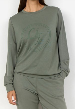 Femme portant une chemise à manches longues vert olive et un pantalon assorti, le texte sur la chemise dit "Life" avec des mots circulaires "Thankful Grateful Blessed."