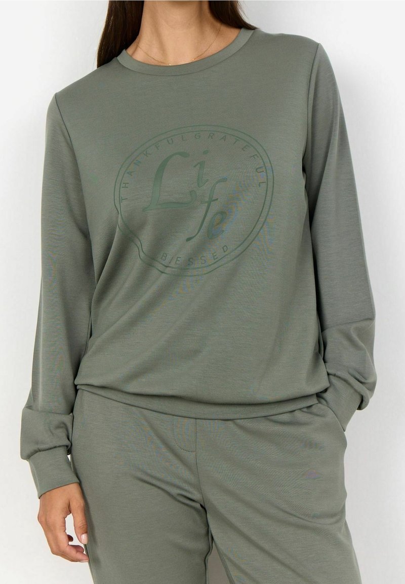 Femme portant une chemise à manches longues vert olive et un pantalon assorti, le texte sur la chemise dit "Life" avec des mots circulaires "Thankful Grateful Blessed."