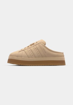 Fløyelsbeige slip-on sneakers med rund tå, tykk platåsåle, strukturert overdel og tre vertikale striper på siden.