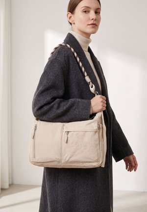 Frau in einem dunkelgrauen Mantel trägt eine beige GMV-Schultertasche mit vorderen Reißverschlusstaschen und einem geflochtenen Riemen über der Schulter im Innenbereich.
