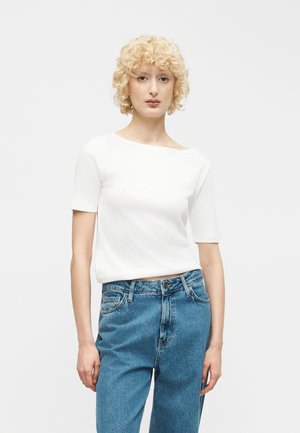 Rövid, göndör, szőke hajú nő, fehér rövid ujjú felsőt és magas derekú kék farmert visel, sima, világos háttér előtt áll.