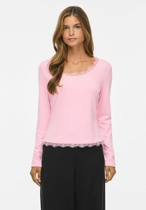 Jeune femme aux longs cheveux bruns portant un haut rose clair à manches longues avec bordure en dentelle et un pantalon noir, debout devant un fond uni.