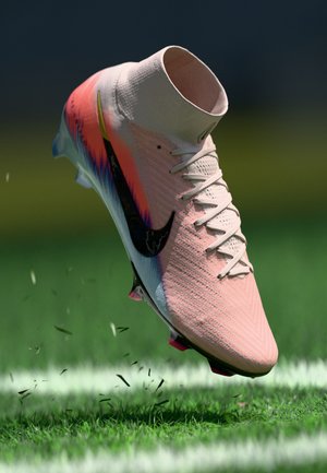 Botín de fútbol Nike rosa y blanco con swoosh negro levantando hierba en un campo verde con líneas blancas.