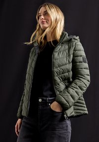 Vadderad olivgrön jacka med huva, prydd med diagonala sömmar, dragkedjefickor och ett texturerat tyg. Svart skjorta och jeans under.