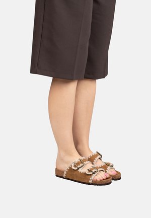 Persona che indossa sandali doppi in camoscio marrone con cuciture bianche e fibbie nere, abbinati a pantaloni scuri corti.